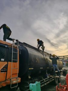 A group of industrial workers climb an oil tanker truck under a moody evening sky.